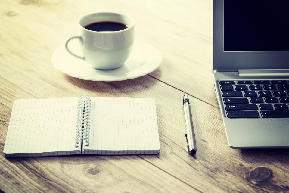 Libreta abierta y pluma junto a una laptop y una taza de café sobre una mesa de madera