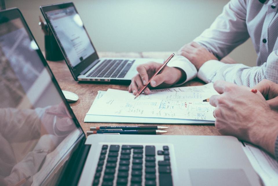 Manos anotando una lista sobre papel junto a dos laptops en una mesa de trabajo