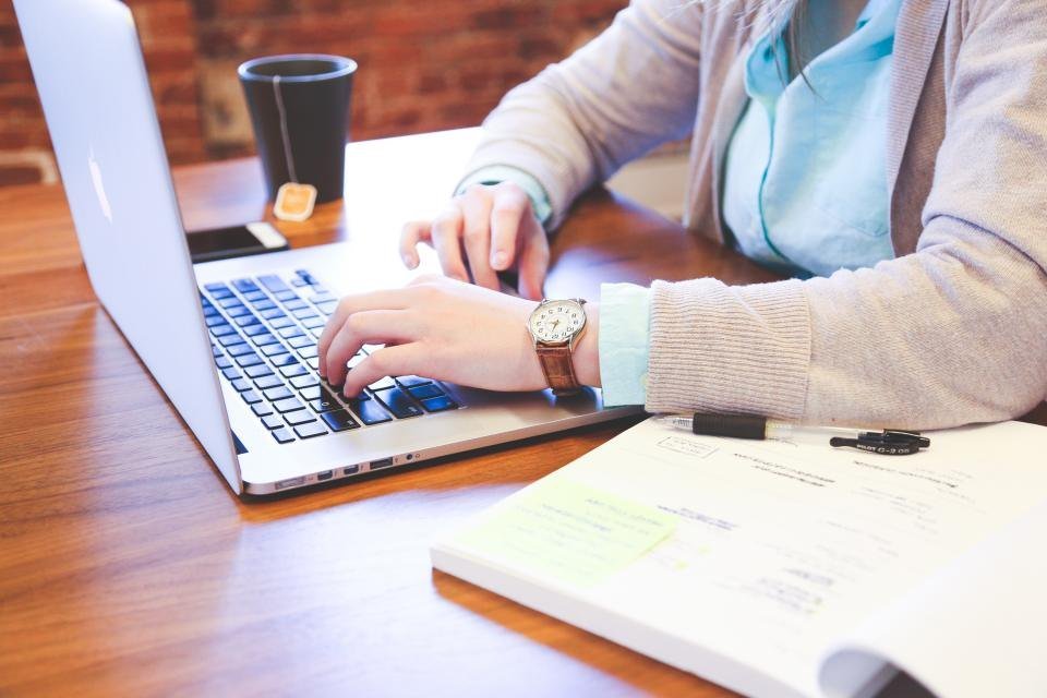 Manos escribiendo en una laptop junto a una libreta con notas
